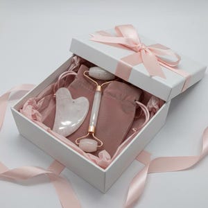 Op de afbeelding: Een witte geschenkdoos, vastgebonden met een roze lint, met een roze fluwelen zakje, een rozenkwarts gezichtsroller en een hartvormige gua sha tool. De doos is gevuld met roze vloeipapier.