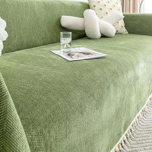 Pode incluir: Uma capa de sofá verde com um padrão de espinha de peixe. A capa tem uma franja com pequenas borlas redondas ao longo da borda. Um copo de água com uma flor e uma revista estão no sofá.