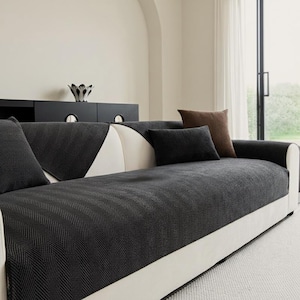 Puede incluir: Un sofá moderno con base blanca y funda con estampado de espiga gris oscuro. El sofá está adornado con varias almohadas en tonos grises y marrones. Una pieza decorativa negra se encuentra sobre un mueble negro en el fondo.