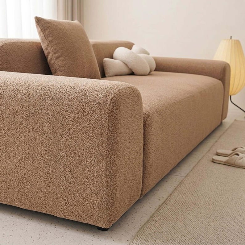 May include: A modern, light brown sofa with a textured, plush fabric. The sofa has a large, square armrest and is accompanied by a matching pillow and a decorative, white, cloud-shaped pillow. The room also features a floor lamp and a rug.
