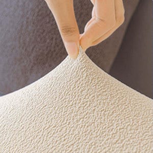 May include: Close-up of a beige textured fabric being pulled up by a hand. The fabric has a rough, bumpy surface and appears to be stretchy. The background is a darker shade of brown.