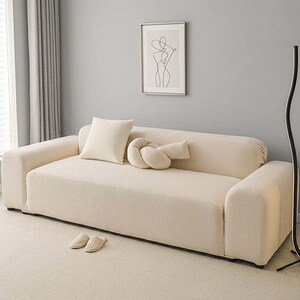 May include: A cream-colored sofa with a matching throw pillow and a decorative knot-shaped pillow. The sofa is in a modern living room setting, with a framed artwork on the wall and a pair of slippers on the floor.