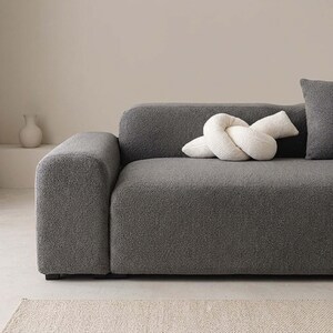 May include: A modern gray sofa with a textured fabric, featuring a rounded armrest and a matching gray pillow. A white knot-shaped decorative pillow rests on the sofa. A small white vase is visible in the background.