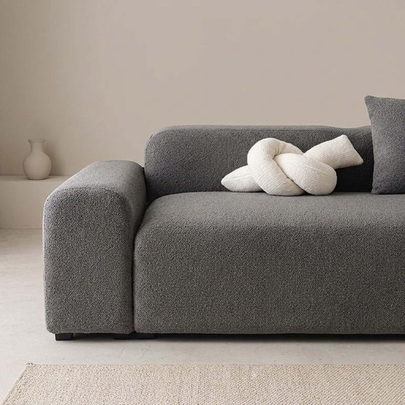 May include: A modern gray sofa with a textured fabric, featuring a rounded armrest and a matching gray pillow. A white knot-shaped decorative pillow rests on the sofa. A small white vase is visible in the background.