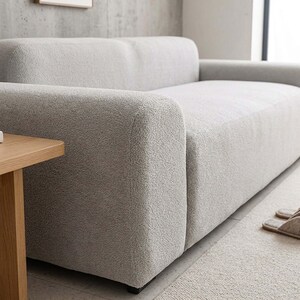 May include: A light gray, modern sofa with a textured, plush fabric. The sofa has a low profile with a rounded armrest and a matching ottoman. A small wooden side table is visible on the left.