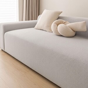 May include: A light gray, textured sofa with two decorative pillows. One pillow is rectangular, and the other is a knotted design. The pillows and sofa are neutral colors, creating a calming aesthetic. The sofa is placed in front of a window with beige curtains.