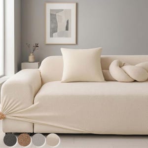 May include: A cream-colored sofa cover is being stretched over a sofa. A square cream-colored pillow and a decorative knotted pillow are on the sofa. The sofa is in front of a gray wall with a framed abstract art print.
