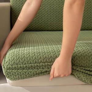 May include: A green sofa cushion cover with a textured leaf pattern. The cover is being placed on a sofa cushion. The cover is a solid green color and appears to be made of a soft, knitted material. The sofa cushion is white.