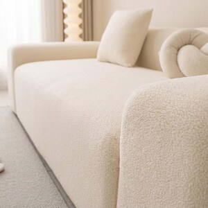May include: Cream-colored, textured sofa with a rounded armrest. The sofa has a matching square pillow and a decorative knot-shaped pillow. The fabric has a soft, plush appearance. The sofa is in a well-lit room.