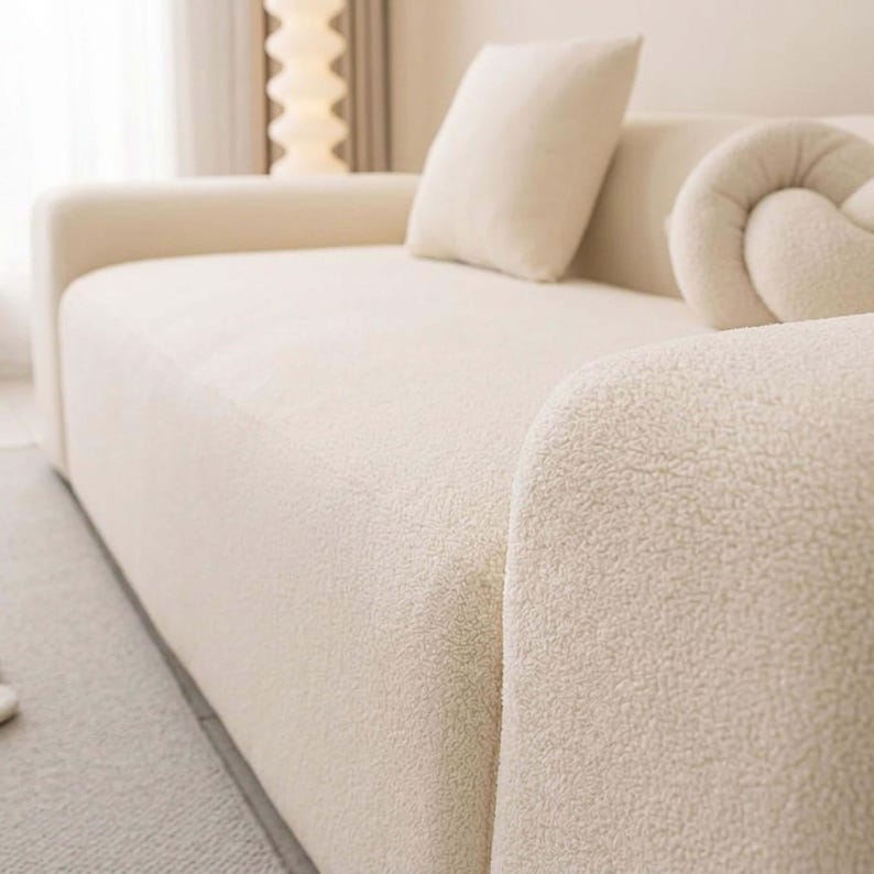 May include: Cream-colored, textured sofa with a rounded armrest. The sofa has a matching square pillow and a decorative knot-shaped pillow. The fabric has a soft, plush appearance. The sofa is in a well-lit room.