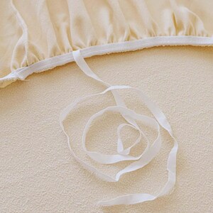 May include: Close-up of a cream-colored fabric with a white elastic band and a white ribbon. The fabric has a soft, textured appearance. The ribbon is loosely coiled, creating a gentle, flowing design.