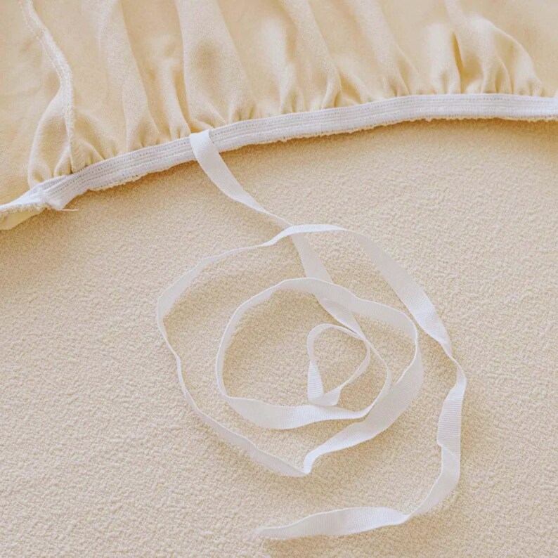 May include: Close-up of a cream-colored fabric with a white elastic band and a white ribbon. The fabric has a soft, textured appearance. The ribbon is loosely coiled, creating a gentle, flowing design.