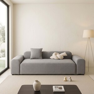 May include: A modern gray sofa with a square gray pillow and a cream-colored knot-shaped pillow. The sofa sits on a beige rug in a well-lit room. A floor lamp and a dark brown coffee table are also visible.