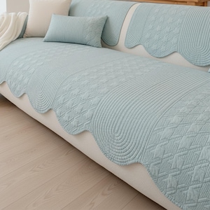 Puede incluir: Una funda de sofá acolchada de color azul claro con borde festoneado. La funda presenta un patrón geométrico y va acompañada de cojines a juego. El sofá está sobre un suelo de madera clara.