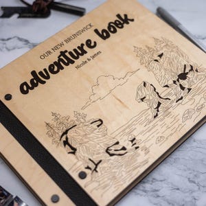 May include: A rectangular wooden photo album with a light wood grain finish. The cover features the engraved text "OUR NEW BRUNSWICK adventure book" and "Nicole & James" above a line art illustration of rocky coastal cliffs and pine trees.