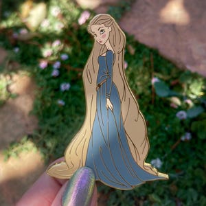 May include: A gold-colored enamel pin featuring a cartoon illustration of a woman with long blonde hair wearing a blue dress. The pin has a detailed design with a smooth finish. The text "@CURSEDCONCEPTS" is at the bottom.