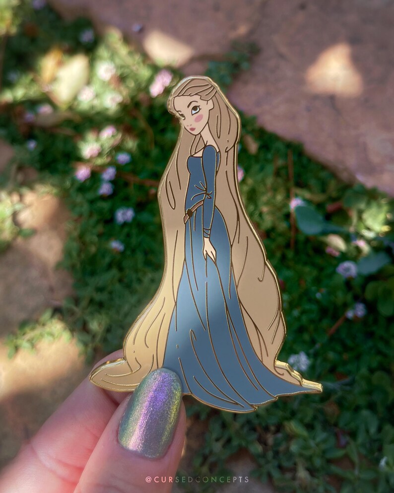 May include: A gold-colored enamel pin featuring a cartoon illustration of a woman with long blonde hair wearing a blue dress. The pin has a detailed design with a smooth finish. The text "@CURSEDCONCEPTS" is at the bottom.