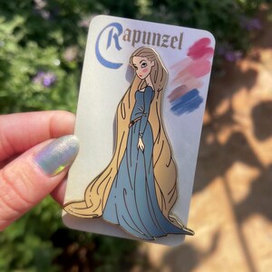 May include: A Rapunzel enamel pin featuring the character in a blue dress with long blonde hair. The pin is on a white card with the name "Rapunzel" and color swatches. The pin is held by a person.