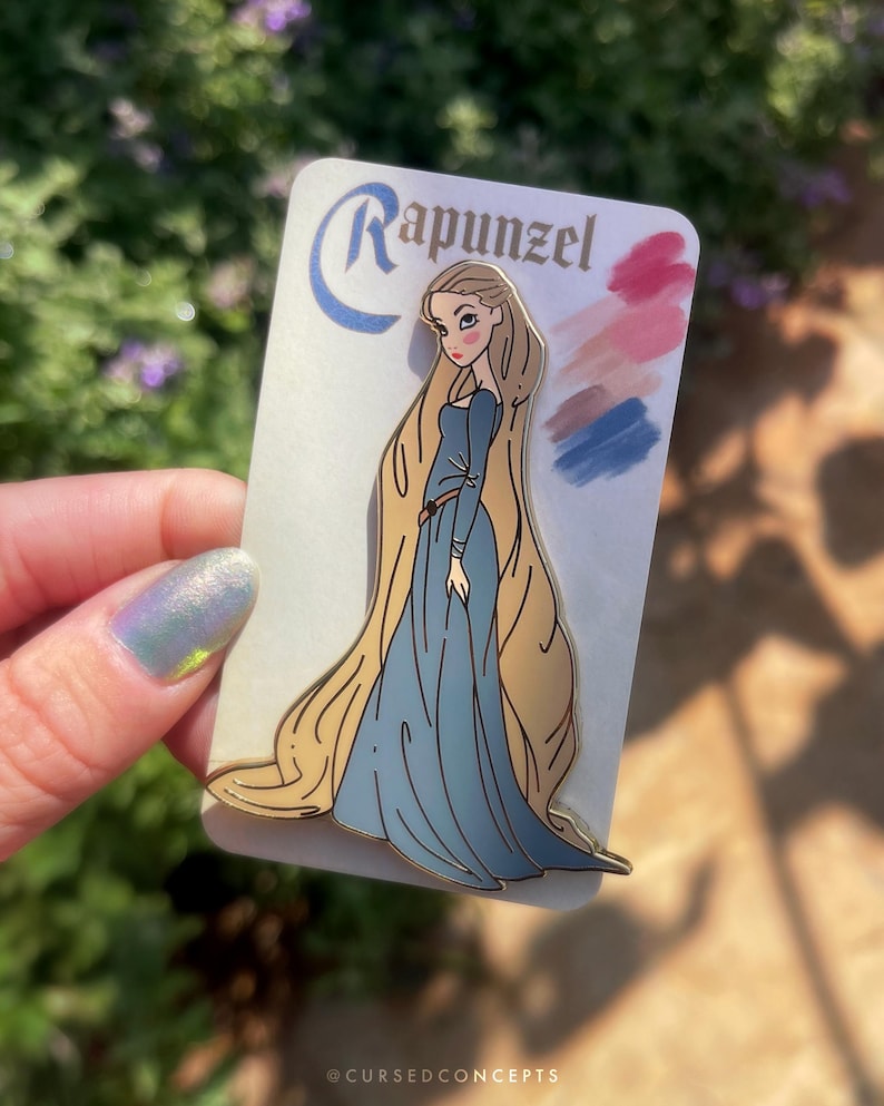 May include: A Rapunzel enamel pin featuring the character in a blue dress with long blonde hair. The pin is on a white card with the name "Rapunzel" and color swatches. The pin is held by a person.