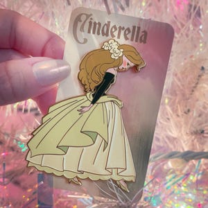 May include: A Cinderella enamel pin featuring the character in a yellow gown, black sleeves, and brown hair. The pin has a gold outline and the word "Cinderella" at the top. The pin is held against a pink and white background.
