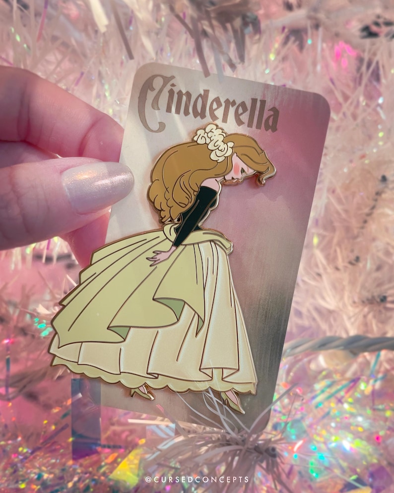 May include: A Cinderella enamel pin featuring the character in a yellow gown, black sleeves, and brown hair. The pin has a gold outline and the word "Cinderella" at the top. The pin is held against a pink and white background.