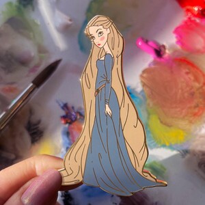 May include: A gold-toned enamel pin featuring a cartoon illustration of a woman with long blonde hair and a blue dress. The pin has a detailed design with a brown belt and is held against a colorful background. The text "@CURSEDCONCEPTS" is at the bottom.