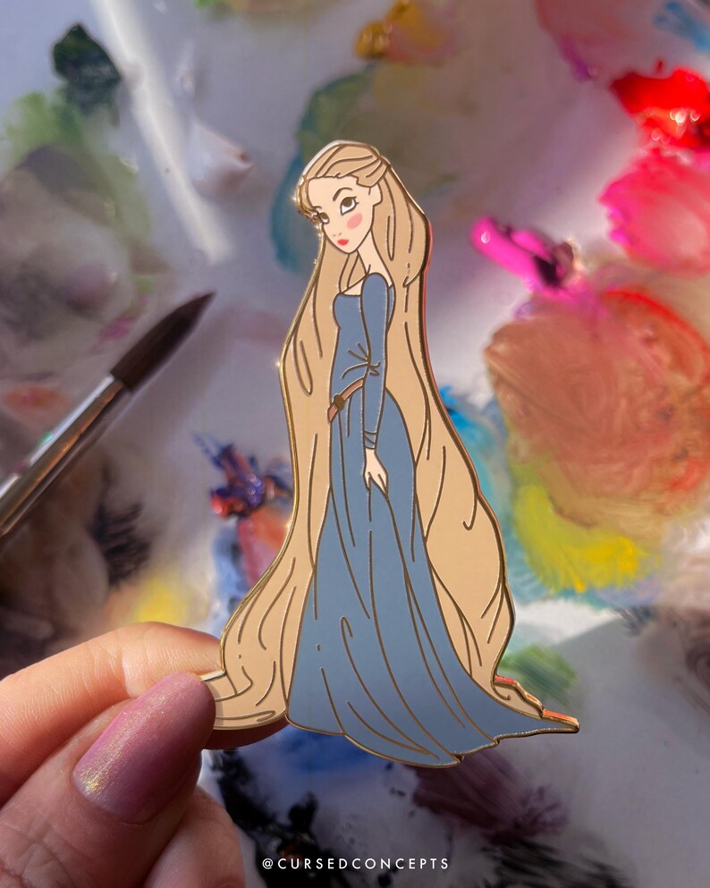 May include: A gold-toned enamel pin featuring a cartoon illustration of a woman with long blonde hair and a blue dress. The pin has a detailed design with a brown belt and is held against a colorful background. The text "@CURSEDCONCEPTS" is at the bottom.