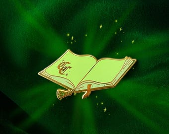 May include: A gold-colored enamel pin in the shape of an open book with light green pages. The book has a gold ribbon bookmark and a gold tassel. The text "CC" is on the left page. The background is a dark green velvet.