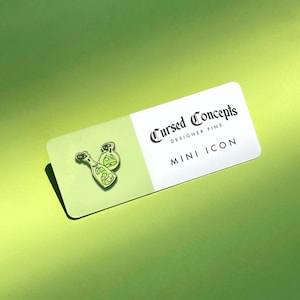 May include: A green and gold enamel pin featuring two bottles with a snake inside. The pin is on a white card with the text "Cursed Concepts Designer Pins Mini Icon".