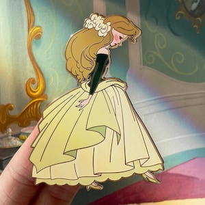 May include: A yellow enamel pin featuring a cartoon character in a yellow gown and a flower in her hair. The character is looking down, with a black top and long blonde hair. The pin has gold-colored outlines and the text "@CURSEDCONCEPTS".