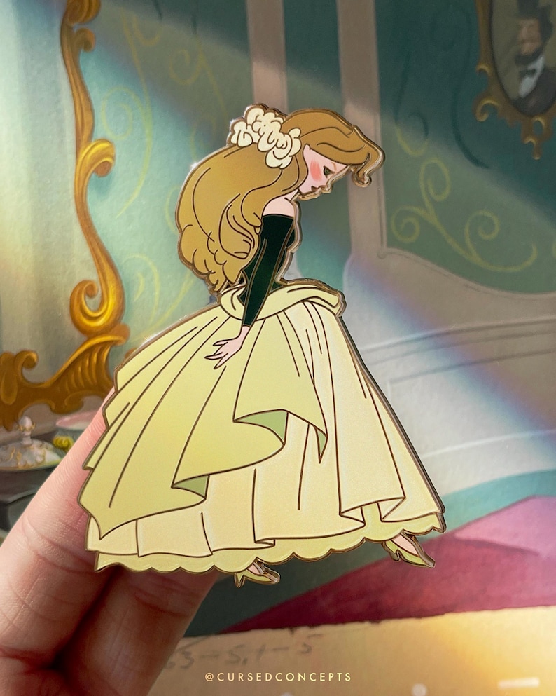 May include: A yellow enamel pin featuring a cartoon character in a yellow gown and a flower in her hair. The character is looking down, with a black top and long blonde hair. The pin has gold-colored outlines and the text "@CURSEDCONCEPTS".