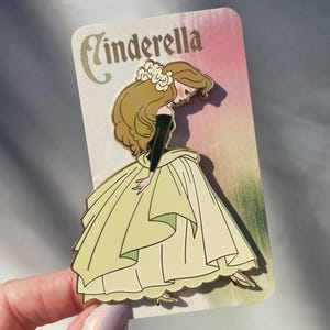 May include: A Cinderella enamel pin featuring the character in a yellow gown with a black top and long blonde hair. The pin is on a card with the word "Cinderella" in gold script. The background is pink and green.
