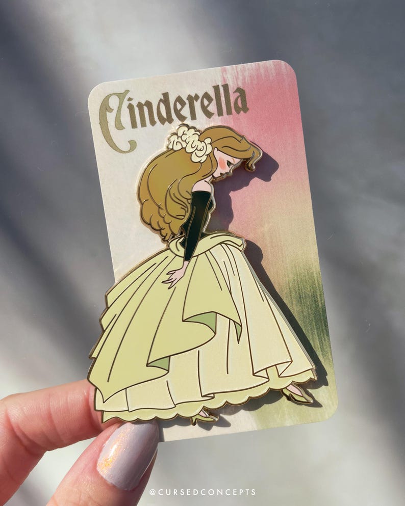 May include: A Cinderella enamel pin featuring the character in a yellow gown with a black top and long blonde hair. The pin is on a card with the word "Cinderella" in gold script. The background is pink and green.
