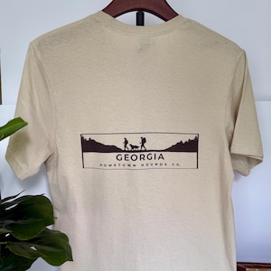 May include: A tan t-shirt featuring a rectangular graphic with the word "GEORGIA" above a silhouette of people hiking with a dog. The text "HOMETOWN HOUNDS CO." is below the graphic. The shirt is hanging on a wooden hanger.