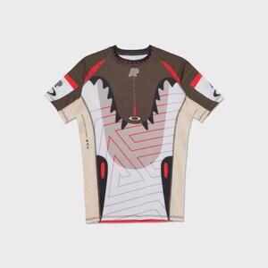 Könnte beinhalten: Ein kurzärmeliges Sportshirt mit einzigartigem Design. Das Shirt hat einen braunen oberen Bereich, weiße und beige Paneele und rote Akzente. Die Vorderseite zeigt ein geometrisches Muster mit dem Oakley-Logo und Text. Das Shirt hat einen Rundhalsausschnitt.