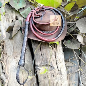 Può includere: Un cappello da cowboy in pelle marrone con una frusta intrecciata e due cordoncini intrecciati neri e rossi. Il cappello ha un logo con la scritta "VAQUERO SPIRIT". Gli oggetti poggiano su una superficie di legno stagionato.