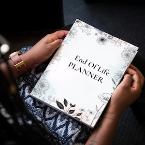 Puede incluir: Un planificador blanco con el texto "End Of Life PLANNER" en negro, sostenido por una persona. La portada presenta un diseño floral en blanco y negro. La persona lleva un reloj con una correa rosa.