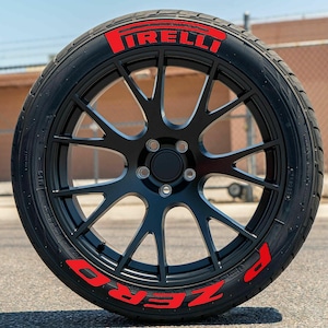 Può includere: Un cerchione nero per auto con uno pneumatico con i loghi rossi "PIRELLI" e "ZERO". Il cerchione ha un design a più razze. L'immagine è ambientata all'aperto.