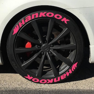 Puede incluir: Primer plano de una rueda de coche negra con un neumático que tiene la palabra "Hankook" en letras de color rosa brillante. La rueda tiene una llanta negra con múltiples radios. Se ve una pinza de freno roja.