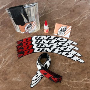 Peut inclure: Un ensemble d'articles de r&eacute;paration de pneus de moto, comprenant des gants noirs, une petite bouteille d'adh&eacute;sif et plusieurs autocollants de pneus Dunlop avec des accents rouges. Un autocollant avec un logo d'ours est &eacute;galement pr&eacute;sent.