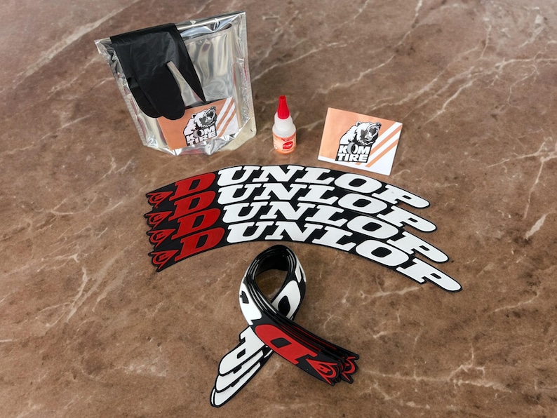 Peut inclure: Un ensemble d'articles de r&eacute;paration de pneus de moto, comprenant des gants noirs, une petite bouteille d'adh&eacute;sif et plusieurs autocollants de pneus Dunlop avec des accents rouges. Un autocollant avec un logo d'ours est &eacute;galement pr&eacute;sent.