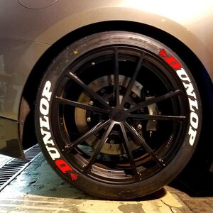 Peut inclure: Gros plan sur un pneu de voiture avec une jante noire et un pneu Dunlop. Le pneu porte l'inscription "DUNLOP" en blanc et des accents rouges. La voiture est de couleur gris fonc&eacute;.
