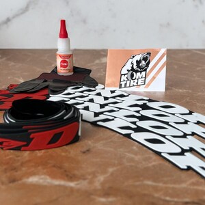 Peut inclure: Ensemble d'accessoires pour pneus de v&eacute;lo, comprenant des protections de jantes noires et rouges, un tube de colle et une carte avec un logo d'ours et le texte "KOM TIRE". Les protections de jantes ont des lettres blanches.