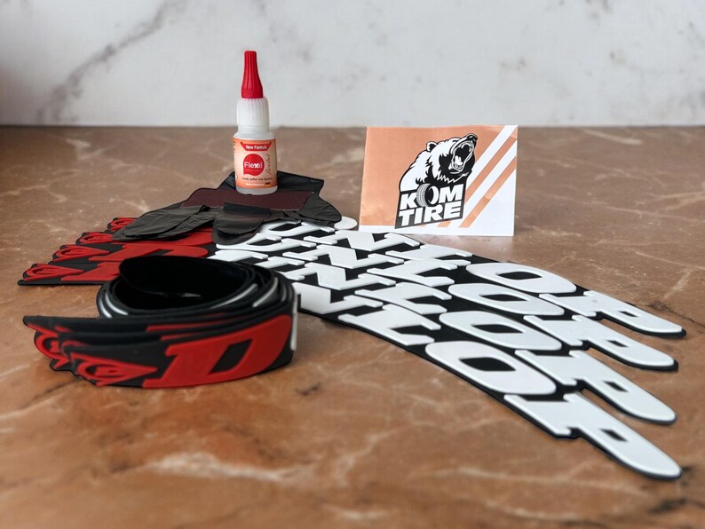 Peut inclure: Ensemble d'accessoires pour pneus de v&eacute;lo, comprenant des protections de jantes noires et rouges, un tube de colle et une carte avec un logo d'ours et le texte "KOM TIRE". Les protections de jantes ont des lettres blanches.