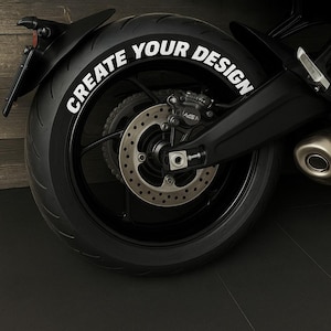 Puede incluir: Primer plano de un neumático de motocicleta negro con las palabras "CREATE YOUR DESIGN" en letras blancas. El neumático está montado en una rueda negra con un freno de disco plateado. El fondo es un panel de madera oscura.