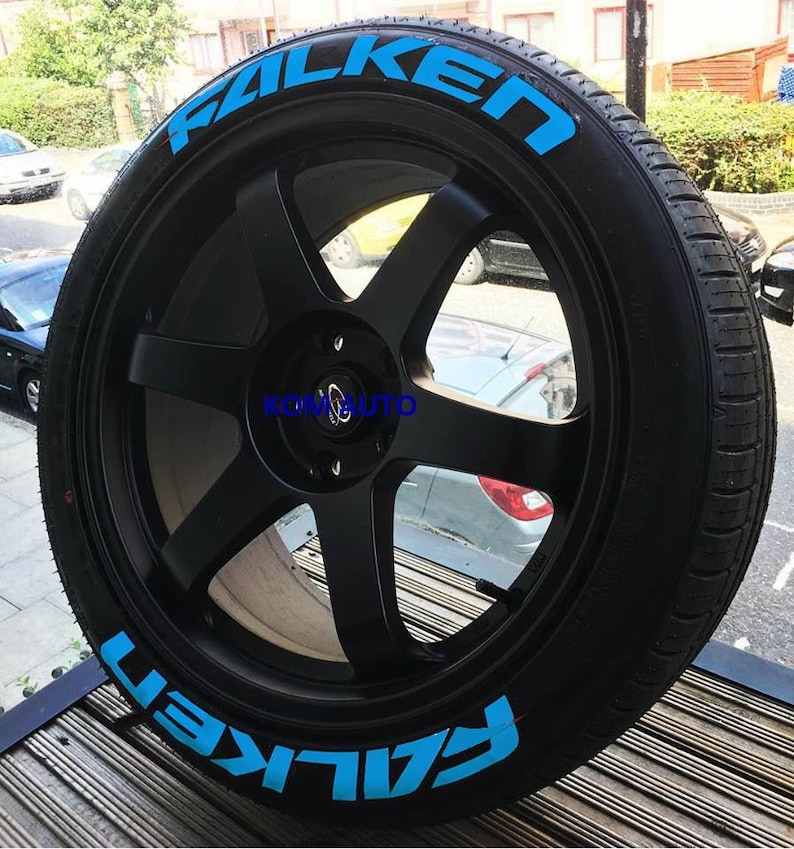 May include: A black car wheel with a tire featuring the word "FALKEN" in bright blue lettering. The wheel has a six-spoke design and a central hubcap. The tire is mounted on a black rim.