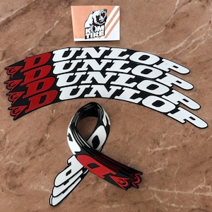 Peut inclure: Ensemble d'autocollants de pneus Dunlop noirs, blancs et rouges. Les autocollants sont incurv&eacute;s et pr&eacute;sentent le logo Dunlop. Un petit autocollant carr&eacute; avec un logo d'ours et le texte "KOM TIRE" est &eacute;galement pr&eacute;sent.