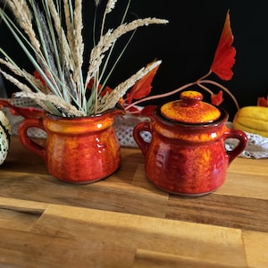 Peut inclure: Deux petits pots en céramique rouge et orange et un pot avec couvercle. L'un des pots contient des herbes séchées. Le pot a un couvercle avec un dessus jaune. Les articles sont sur une surface en bois.