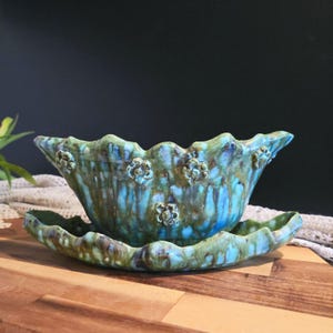 May include: A ceramic planter and saucer set, hand-glazed in shades of turquoise, blue, and green. The planter has a scalloped edge and floral details. The set is designed for displaying plants and has a glossy finish.
