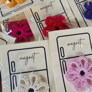 Könnte beinhalten: Eine Sammlung handgefertigter Häkelblumenmagnete in verschiedenen Farben, darunter Gelb, Rot, Rosa, Weiß und Lila. Jeder Magnet ist auf einer Karte mit einer Kühlschrankillustration und dem Wort "Magnet" in Schreibschrift verpackt.
